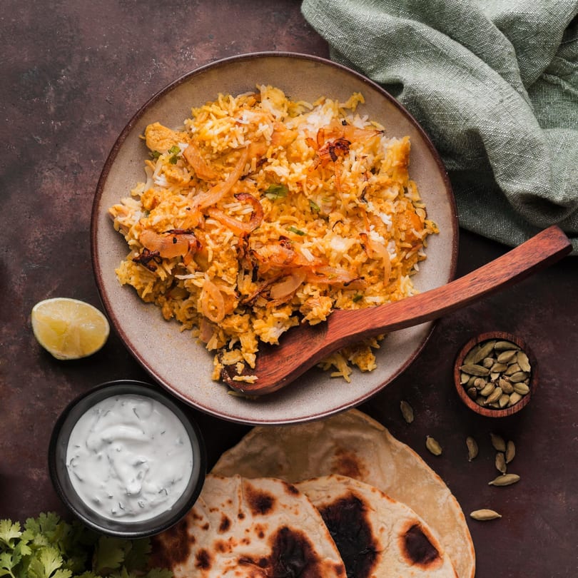 Chicken biryani with fried onions, a side of raita, naan, lime, and cardamom pods.