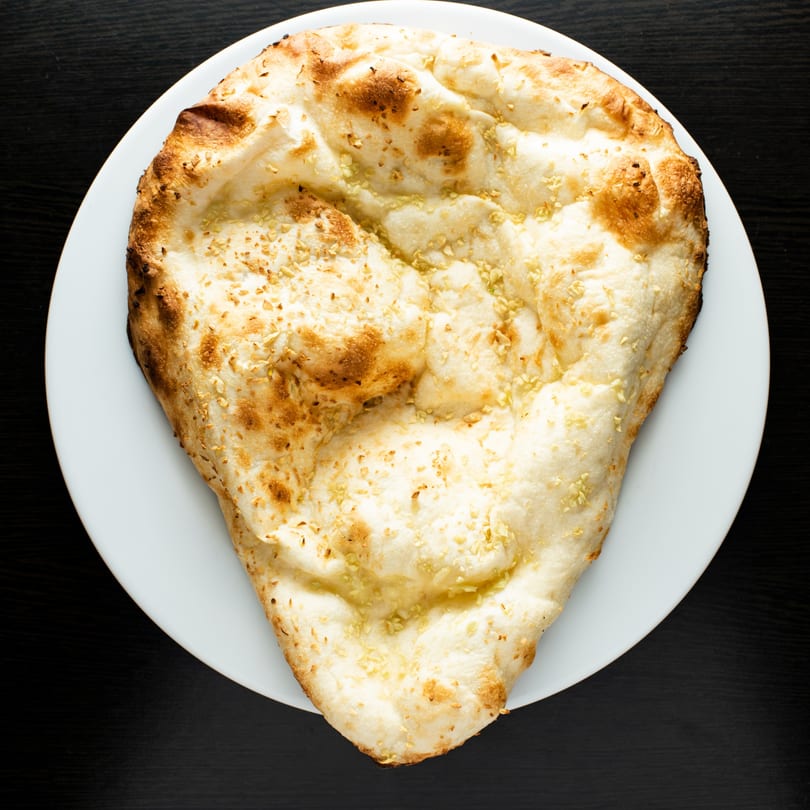 A large, golden-brown garlic naan bread, sprinkled with sesame seeds, on a white plate.