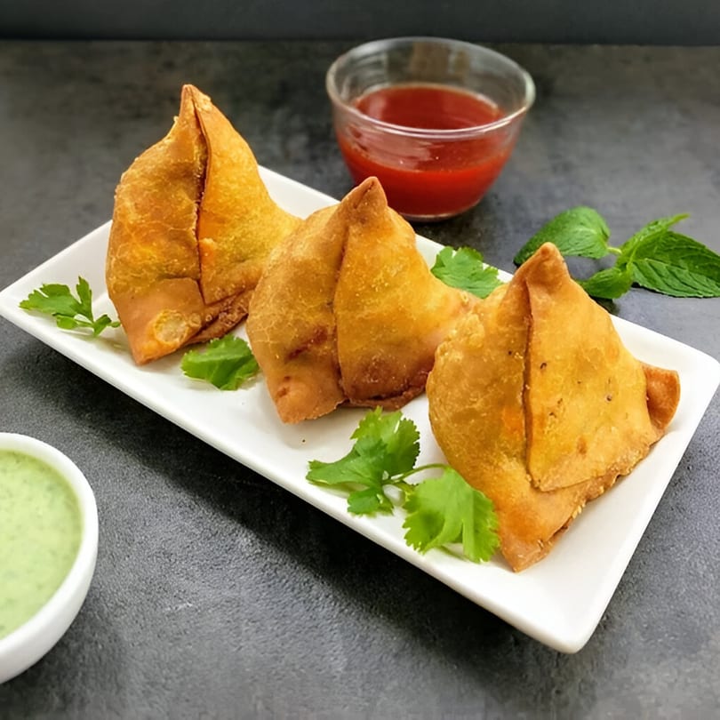 Three golden-brown samosas garnished with cilantro and mint, served with red and green dipping sauces.