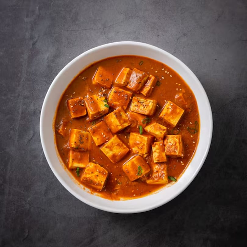 A white bowl filled with paneer curry, featuring golden paneer cubes in a rich orange sauce, garnished with seeds.